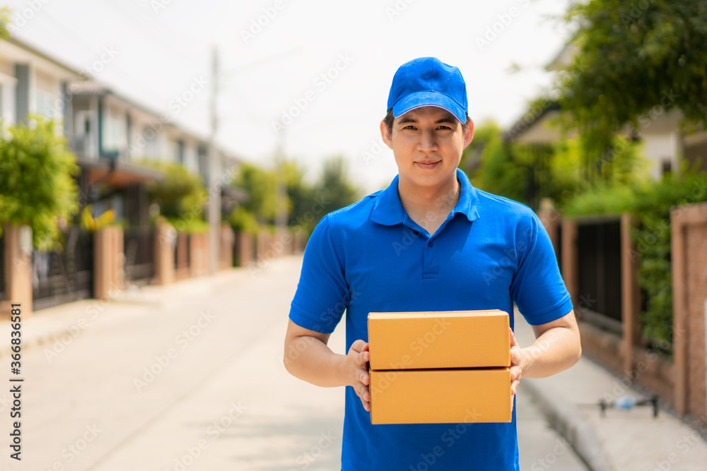 Asian delivery young man in blue uniform smile and holding pile of cardboard boxes in front house vi