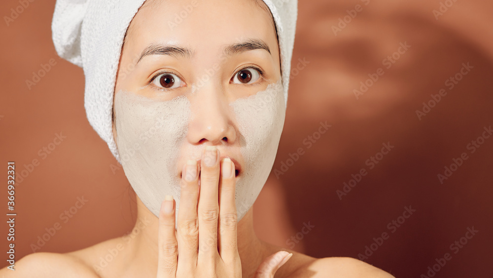 Happy young Asian woman applying clay mask on her face over orange background. Close up.