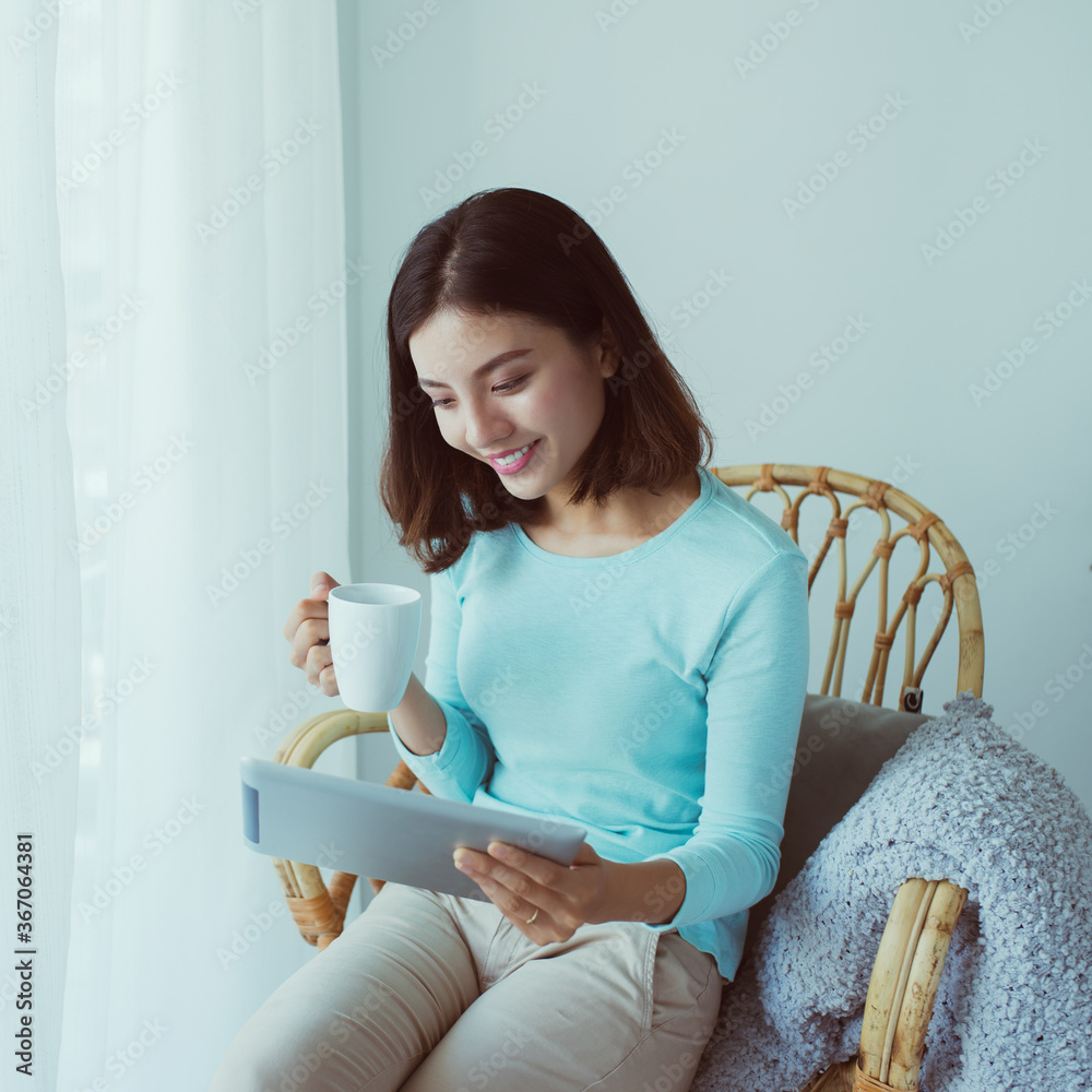 漂亮的年轻亚洲女人正在用ipad，在窗户后面喝咖啡。