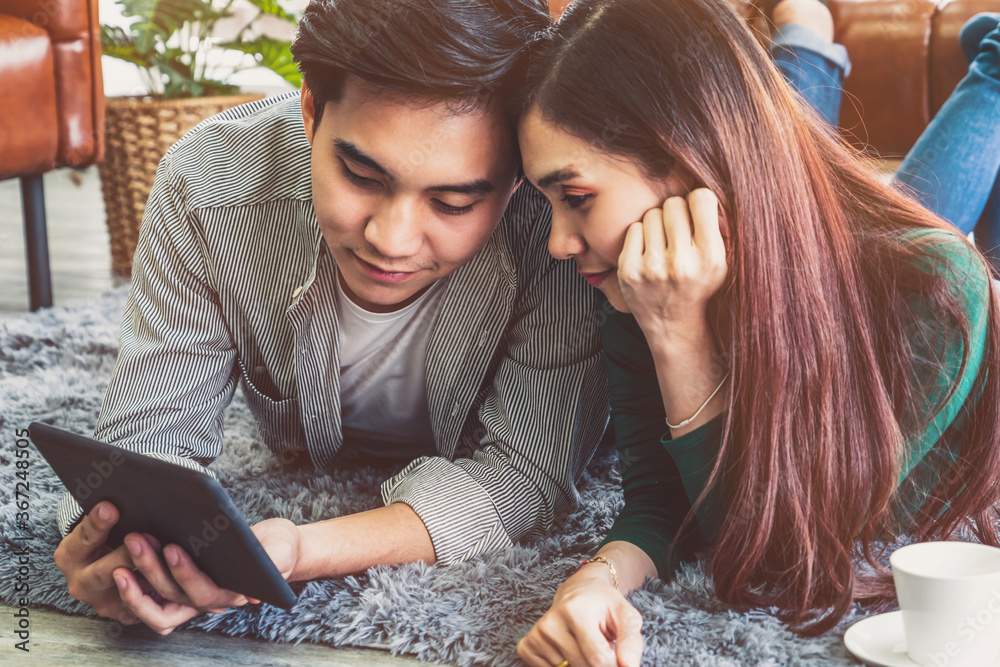 Happy Asian couple use tablet while lying down on carpet at living room floor. Love and relationship