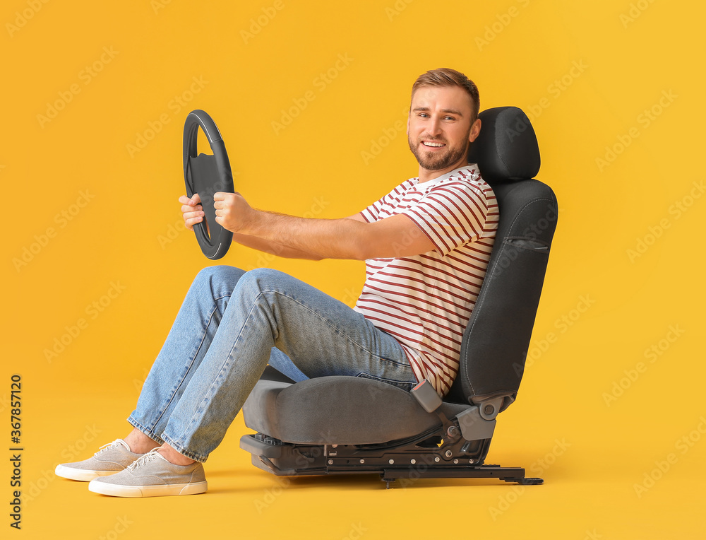 Young man with steering wheel sitting on car seat against color background