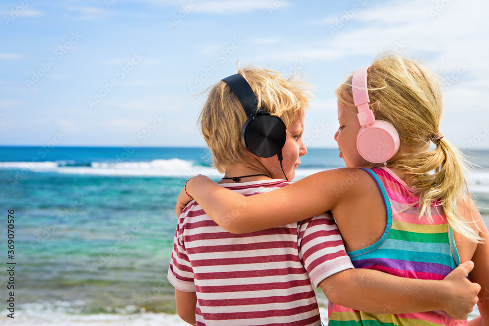 Young positive kids in headphones listening music with fun at tropical beach party. Travel family li