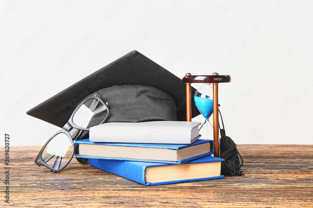 Graduation hat, books and hourglass on table against white background