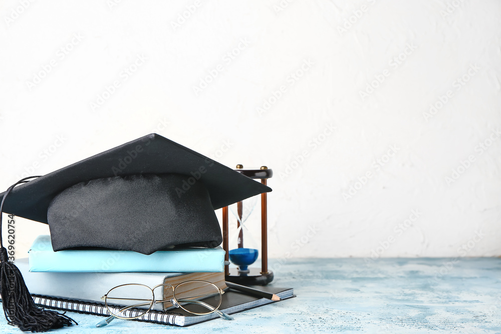 Graduation hat, books and hourglass on table