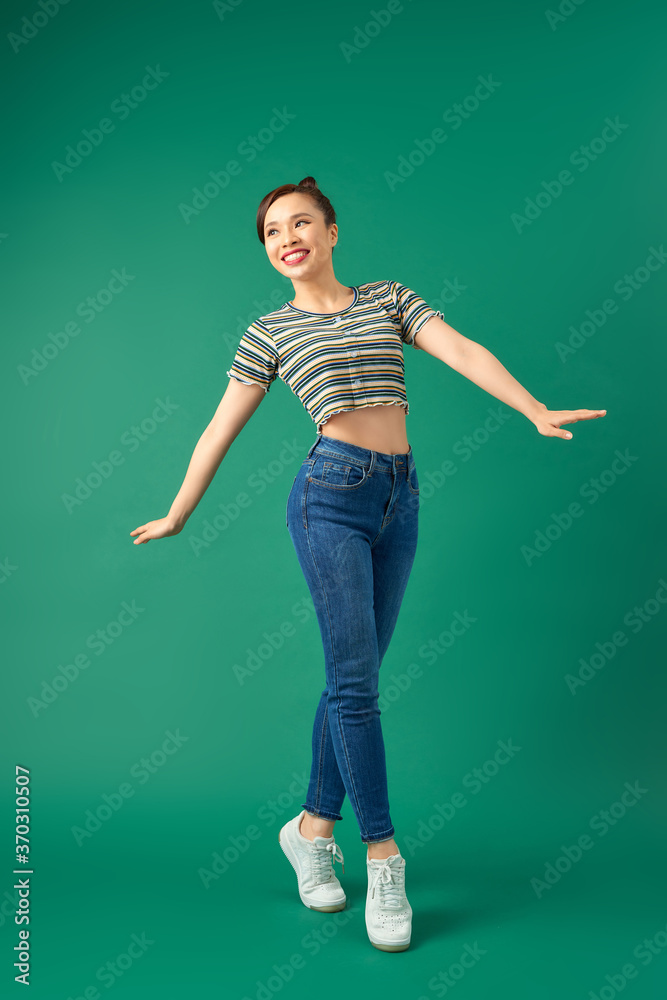 Joyful young Asian woman standing on one leg. Full length view of blissful girl dancing on green bac