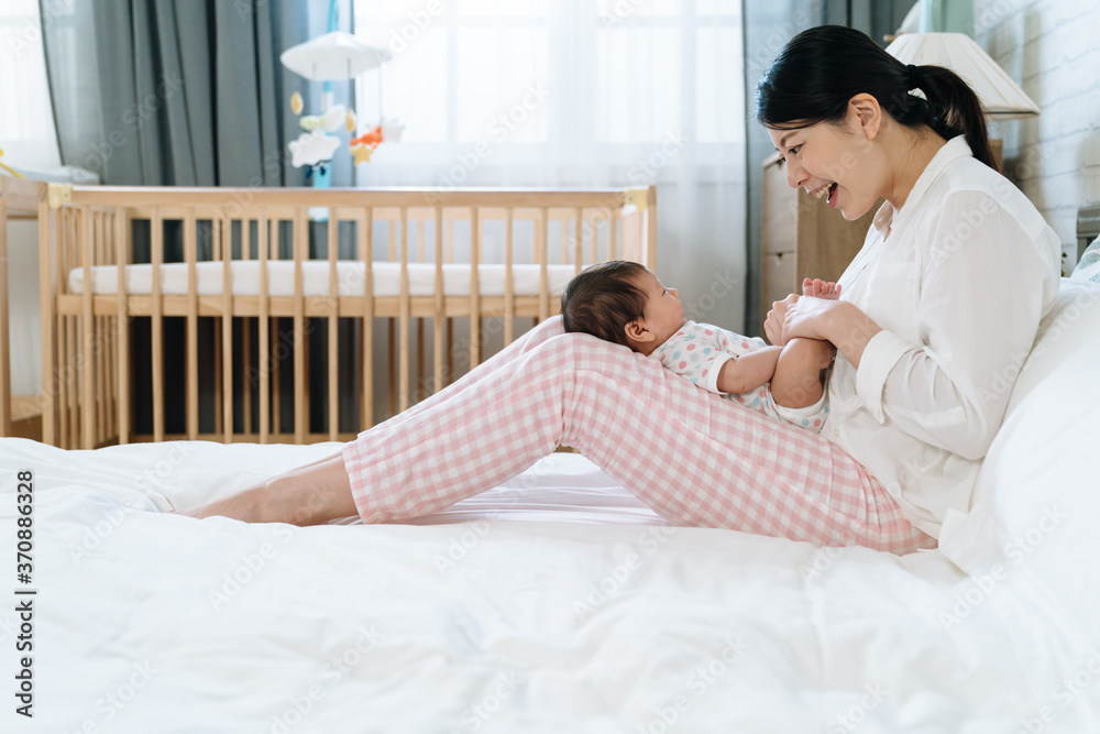 side view of asian new mother talking with joy to her baby whos lying on her legs. chinese mom inte