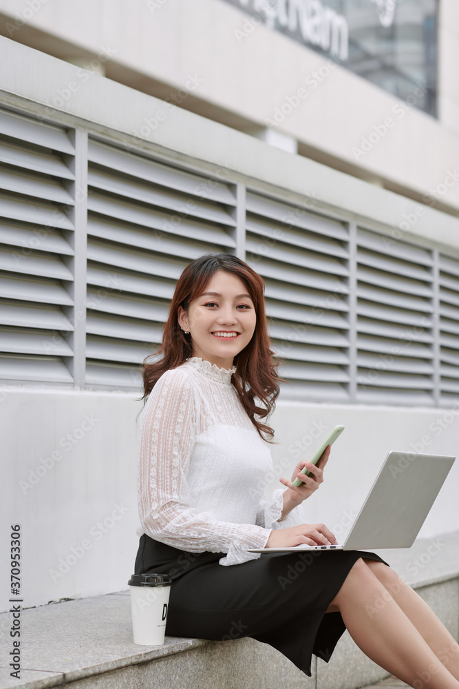 Beautiful business woman sitting on the footsteps in the financial city, using a laptop computer, th