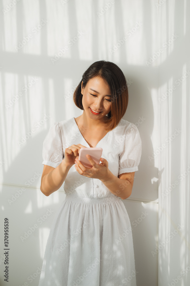 Content woman chatting on mobile phone while standing against white wall in sunset light