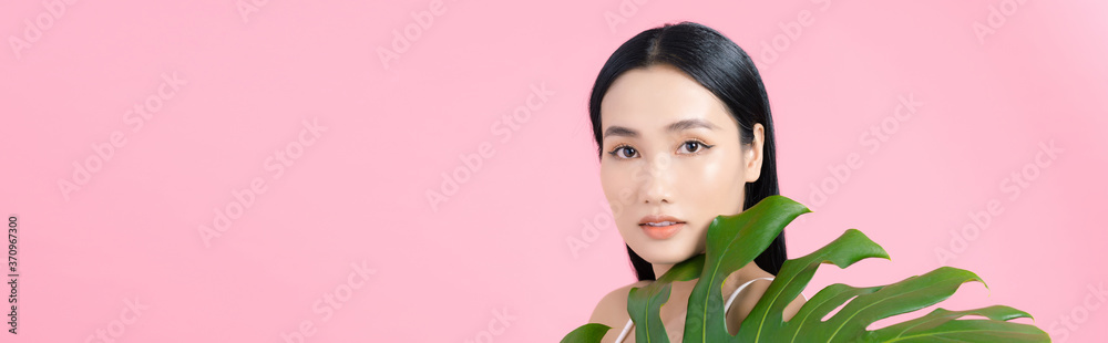 Portrait of young beautiful Asian woman holding tropical leaf on pink background. Natural beauty con