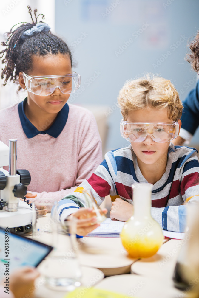 Elementary School Science Classroom: Cute Little Boy in Safety Glasses Mixes Chemicals in Beakers. C