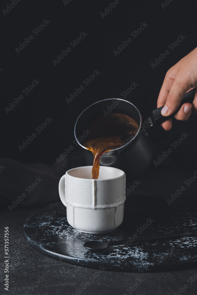 Woman pouring hot coffee from pot into cup on dark background