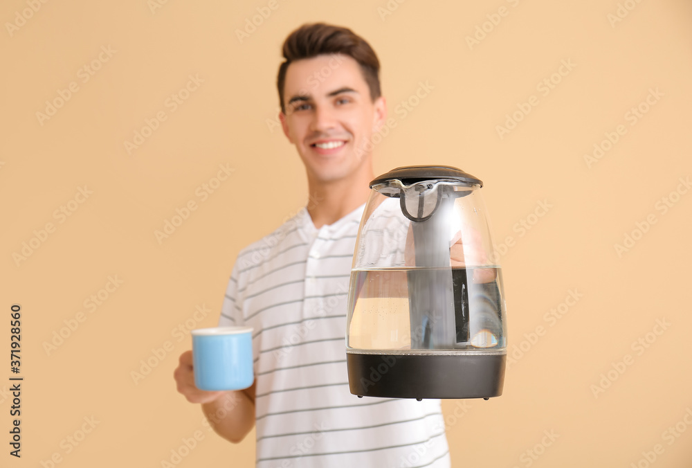 Young man with electric kettle and cup on color background