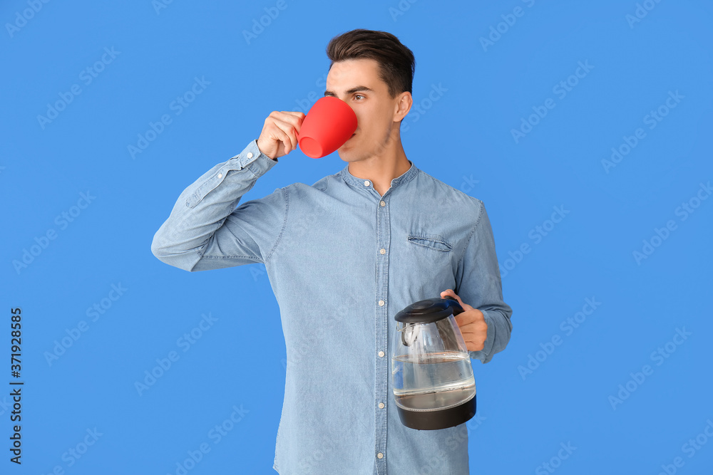 Young man with electric kettle drinking coffee on color background