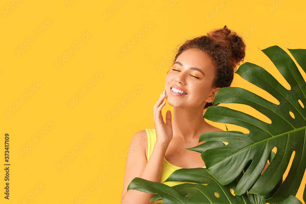 Young African-American woman with beautiful eyeshadows and tropical leaves on color background