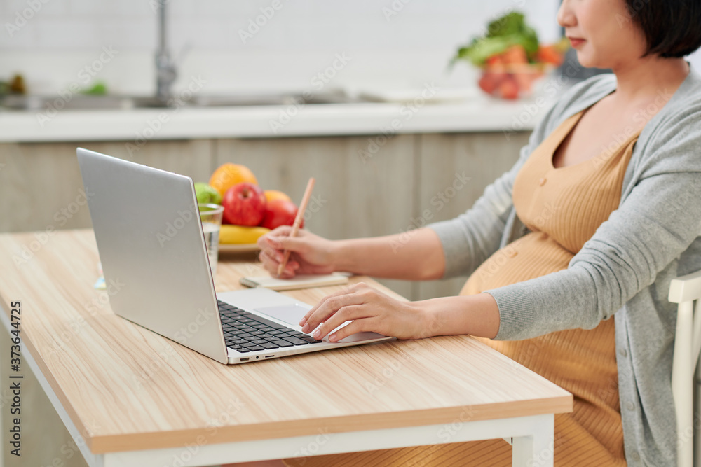 young pregnant woman using laptop while leaning on kitchen counter at home