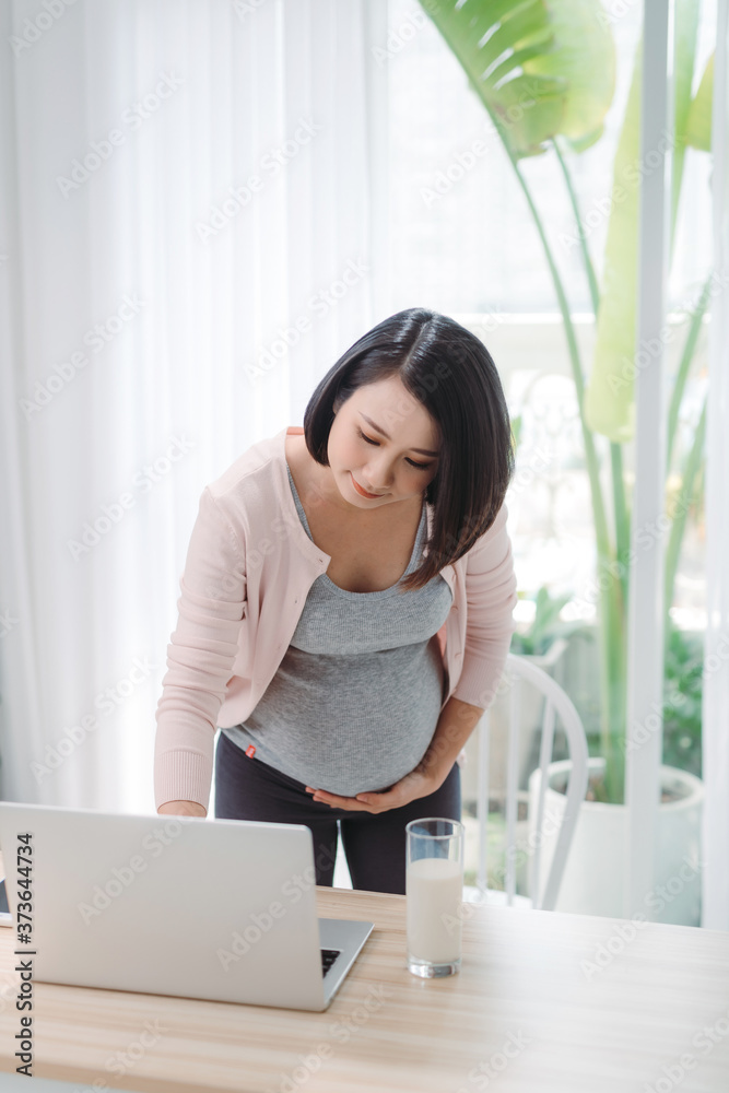 beauty happy Asian pregnant woman with big belly holding glass of milk using computer