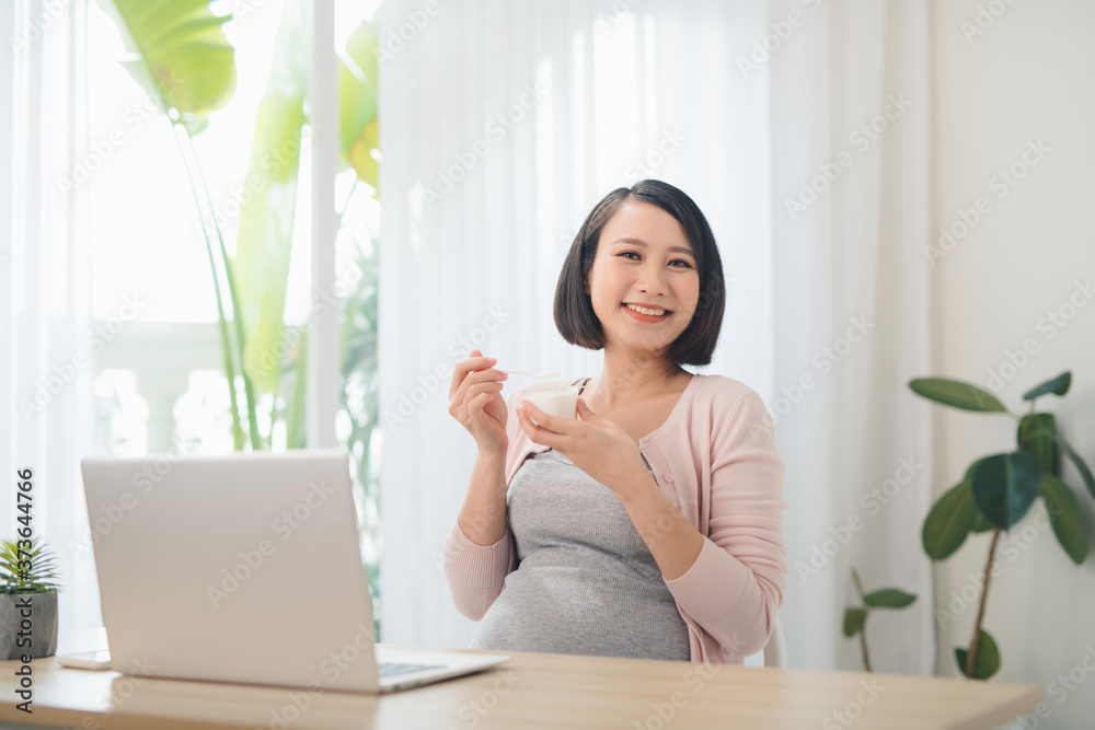 一位面带微笑的红发孕妇在家里拿着笔记本电脑坐在桌子旁吃酸奶