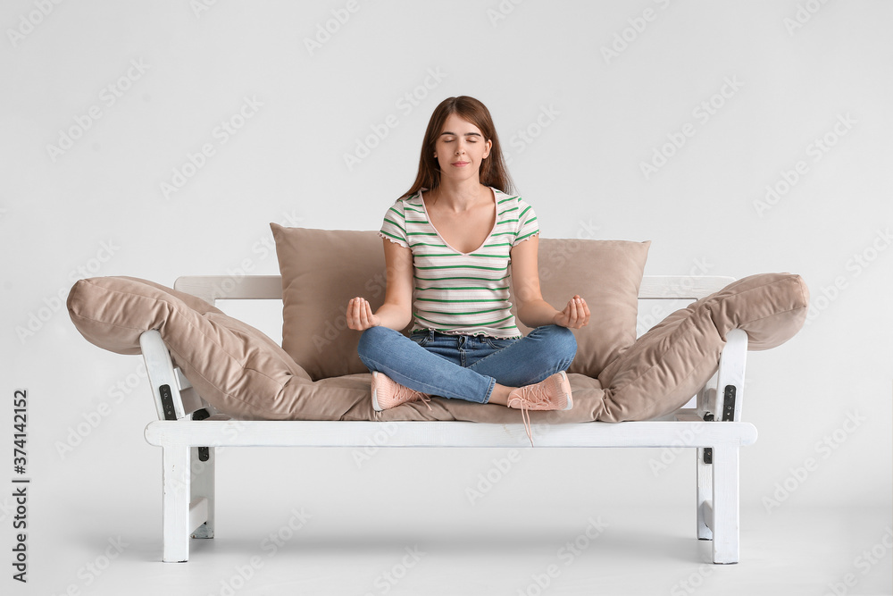 Young woman meditating on modern sofa against light background