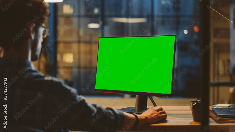 Over the Shoulder:Creative Young Man Sitting at His Desk Using Desktop Computer with Mock-up Green（肩