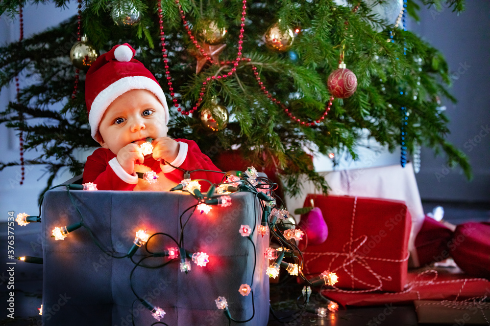 Little cute baby boy in Santa Claus hat holding illuminated lamps sitting in box under Christmas tre