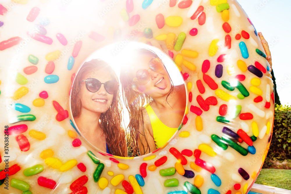 Two teenagers girls stick out tongues posing inside inflatable doughnut wearing sunglasses and smile