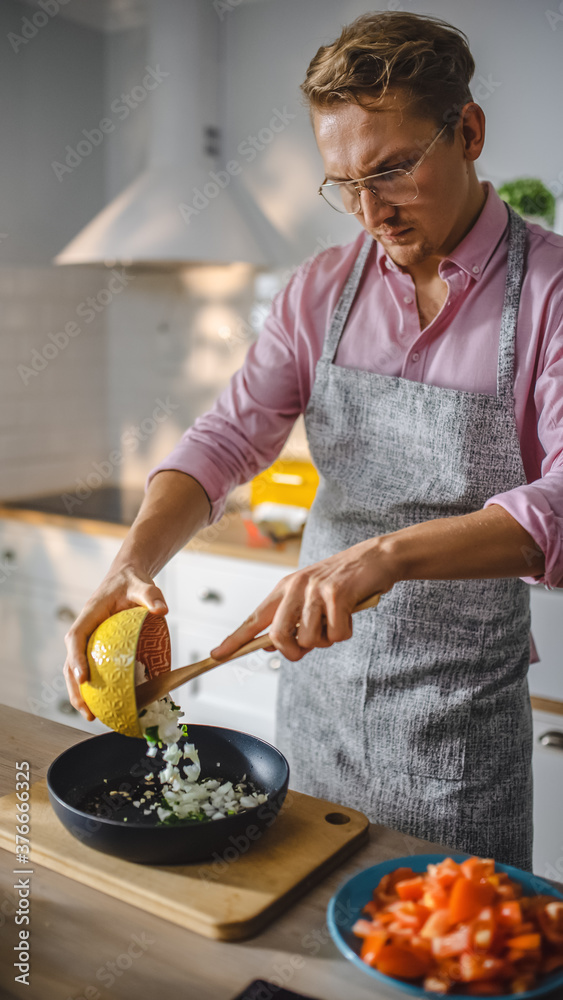 穿着粉色衬衫和围裙的英俊男子正在煎锅上准备健康的素食餐。健康