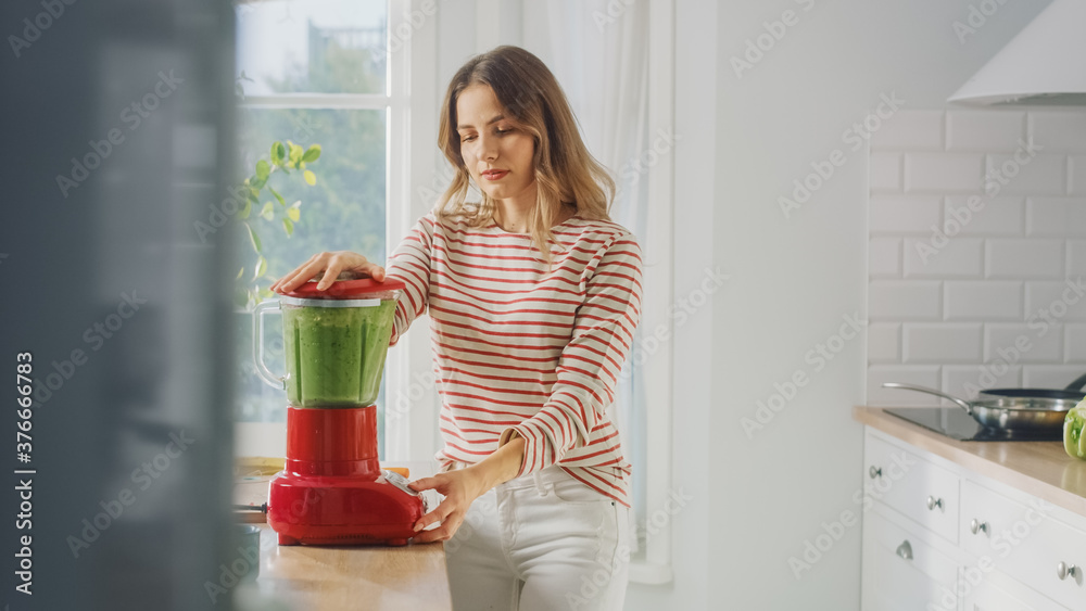 美丽的年轻女性在搅拌机中准备健康的绿色冰沙。正宗的时尚厨房