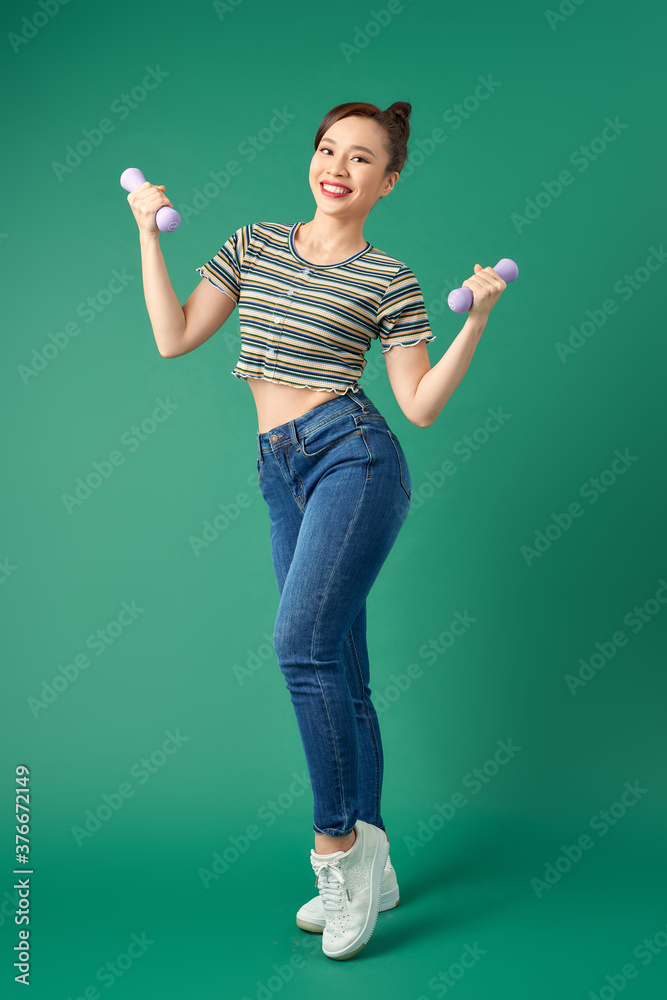 Attractive young Asian woman holding dumbell while standing over green background. Full length.