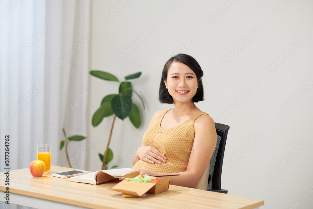 怀孕的年轻女子坐在客厅的桌子上使用数字平板电脑，吃早餐