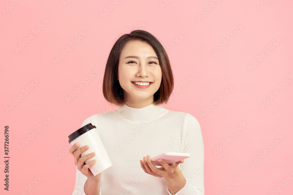 粉色背景下，一位年轻的亚洲女性拿着咖啡杯和智能手机的特写。