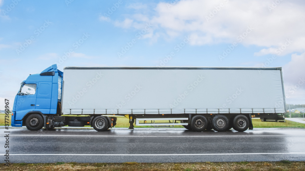 Side View Shot of a Blue Long Haul Semi-Truck with Cargo Trailer Attached Stopped on a Road in the R