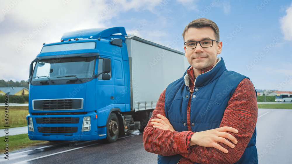 Happy Professional Young Truck Driver Crosses Arms and Smiles on Camera. Behind Him Parked Blue Long