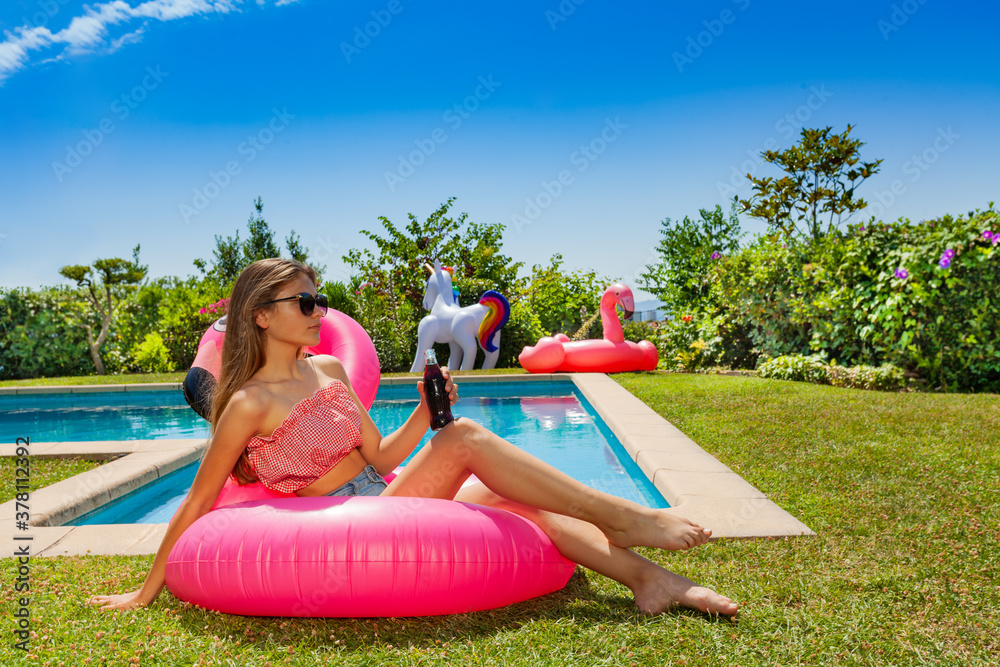 Beautiful girl with soda in hand and sun glasses lay on the pink flamingo near swimming pool in the 