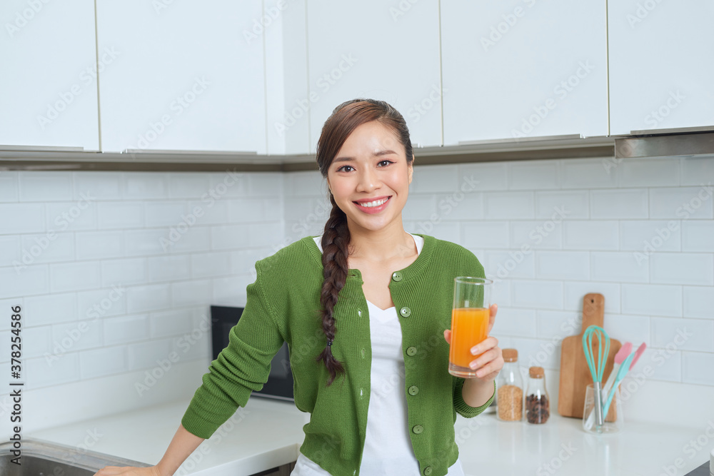 Gorgeous woman drinking a glass of orange juice standing in her kitchen looking at camera