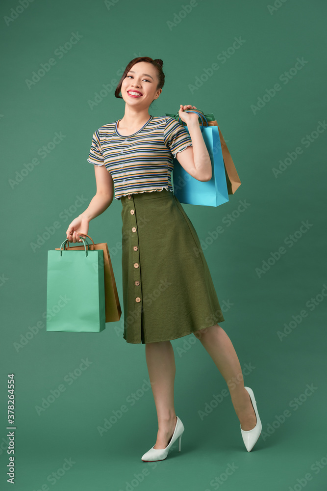 full length view of cheerful, fashionable woman holding shopping bags and smiling on green backgroun