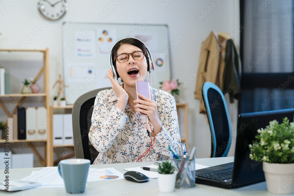 戴着耳机的年轻女时装设计师坐在办公桌旁，惊讶地用手机唱歌