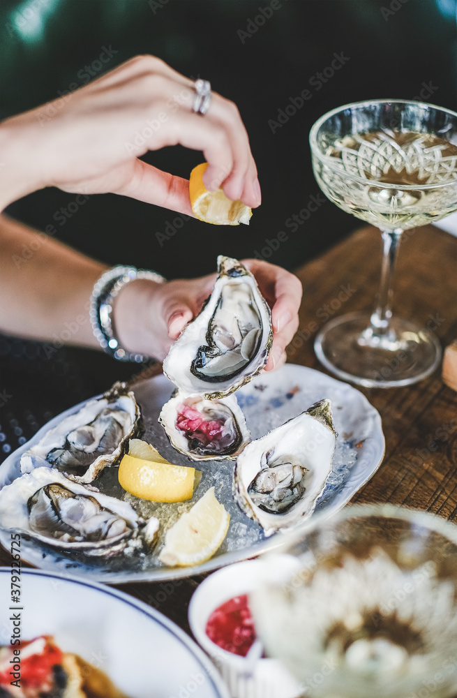 Hands of woman squeezing lemon juice to Irish oysters in plate with ice over glass of champagne in f