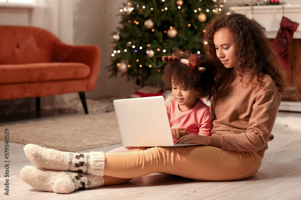 Cute African-American girl and her mother with laptop at home on Christmas eve