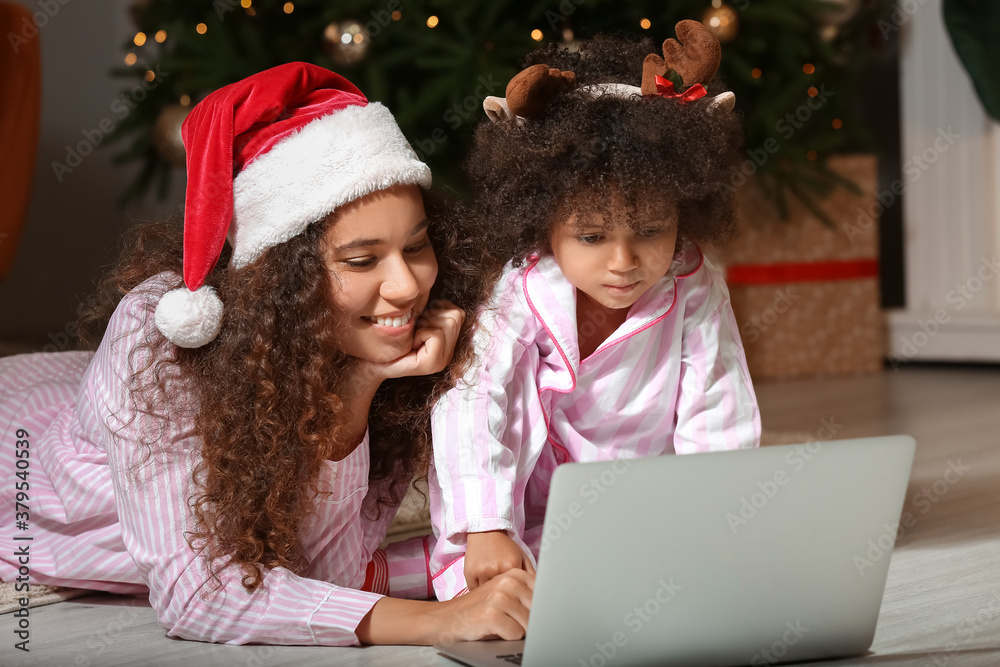 Cute African-American girl and her mother with laptop at home on Christmas eve