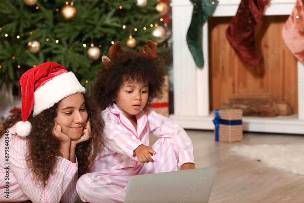 Cute African-American girl and her mother with laptop at home on Christmas eve