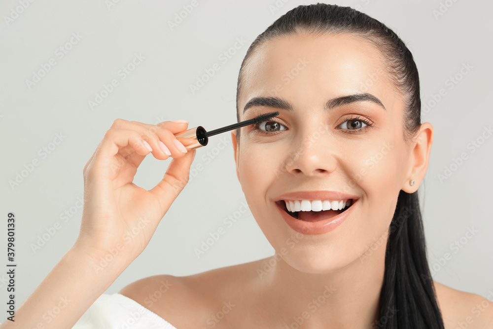 Beautiful young woman applying mascara against light background