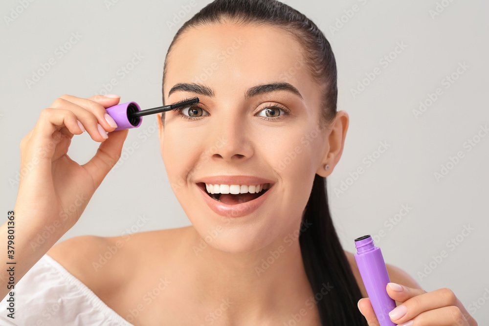 Beautiful young woman applying mascara against light background