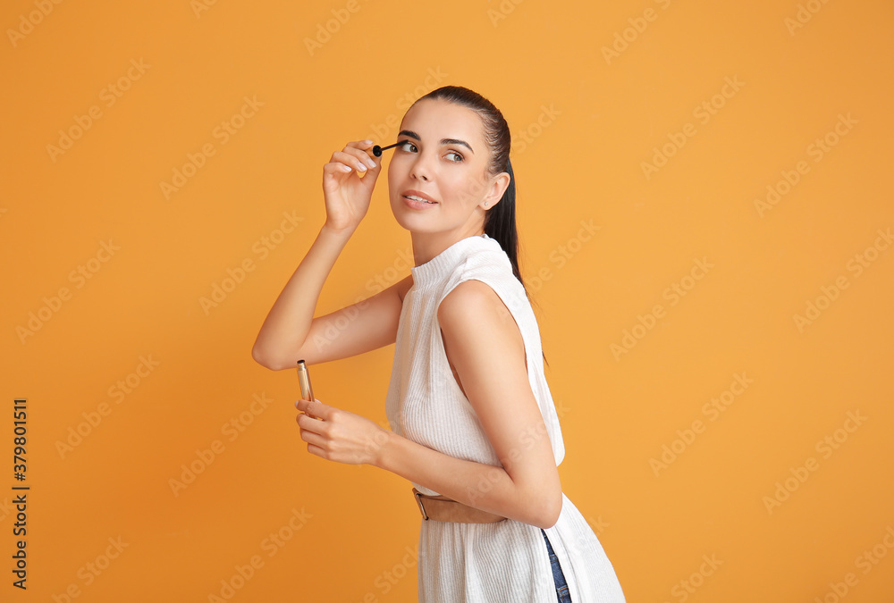 Beautiful young woman applying mascara on color background