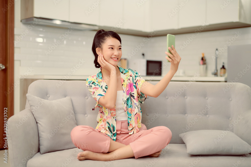 Woman sitting sofa home phone holding hand makes selfie photographs broadcasting smile positive