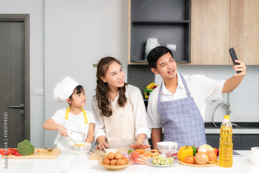 Young Asian family are preparing salad in the kitchen and father take a photo selfie by phone. Excit