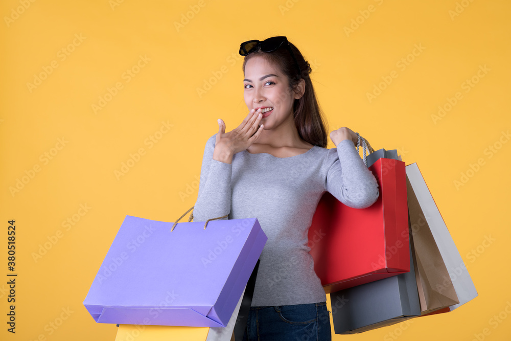 Beautiful young Asian woman carrying shopping bags looking surprised and happy isolated on yellow ba