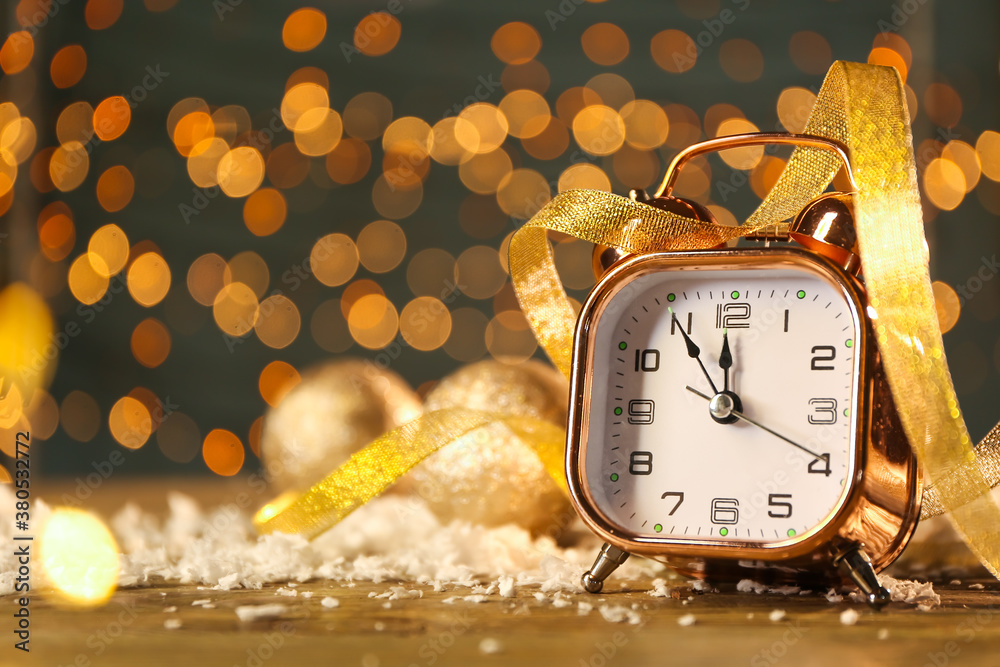 Alarm clock with ribbon and snow on table against blurred lights. Christmas time