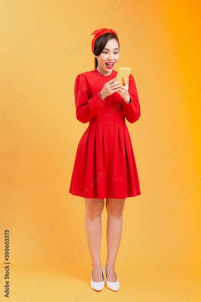 Full length body of surprised young Asian woman reading news while standing over orange background.