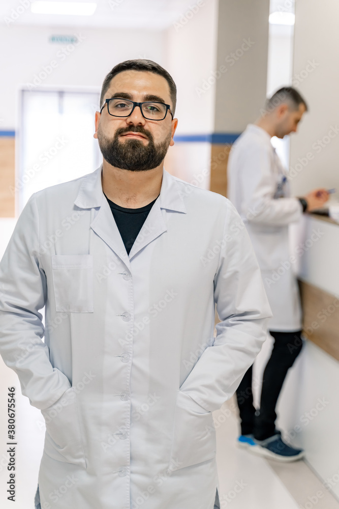 Doctor stands in hospital corridor. Bearded medic in glasses wears scrubs. Health care concept.