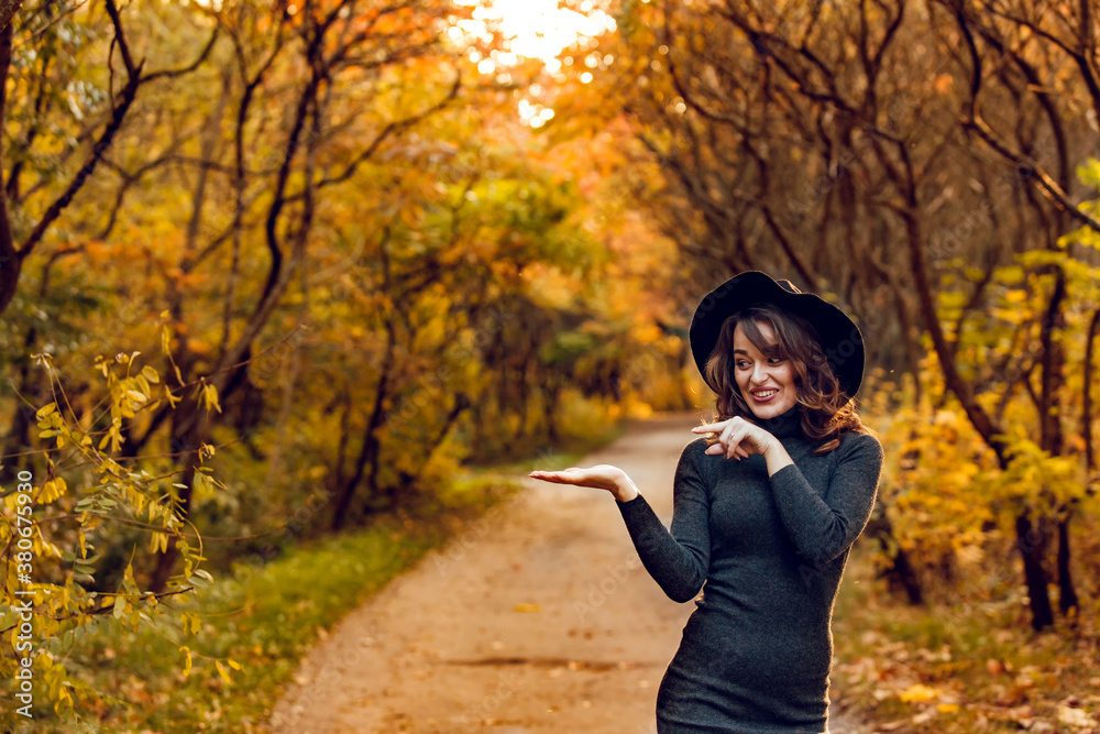 Funny woman in a black hat and a green dress stands in the park. Yellow trees on the background of a